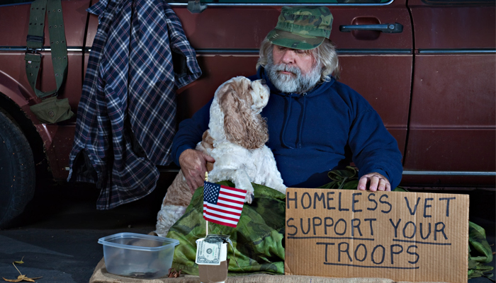 Homeless veteran with his dog holding a “Support Your Troops” sign and donation container beside a truck.