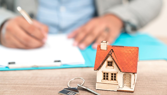 Model house and key on table with person signing paperwork.